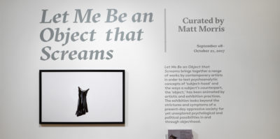 An installation photograph of a title wall in large, grey, bolded text. The text is printed in vinyl and is attached to the wall and writes, “Let Me Be an Object that Screams”. Below the title is an image of a black dress on white paper. The image has a thin, black frame. To the right of the image and title, a paragraph of smaller, bolded text, below a line that writes, “Curated by Matt Morris”.