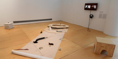 An installation photograph of the corner gallery space. Positioned on the floor, there is a translucent paper about two feet wide and eight feet vertically. Multiple found objects are placed on the translucent paper. A wooden circle protrudes from the ground at the top end of the translucent paper. A television monitor with headphones is mounted to the right wall. On the television monitor there is a middle aged person sitting.