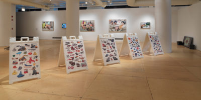 An installation photograph of the gallery space. Situated in the middle of the floor, there are four, white, plastic, A-Frame holders. Each holder is covered with sticker-like designs of assorted objects.