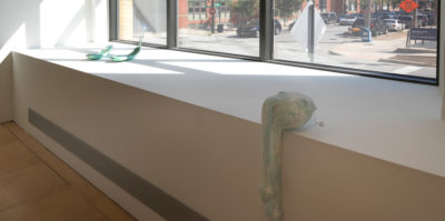 An installation photograph of two objects situated on a wall under a window. The first, closest object is a sculpture that is in the form of a three-dimensional, light blue ball. The shape is situated on the corner of the wall with a drooping shaped accent that hangs below the corner. The second object to the left is of two teal, three-dimensional, oval shaped objects. The oval objects fold in on all sides like a spoon.