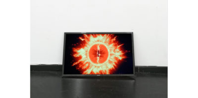 A photograph of a television monitor situated on the floor, against a wall. In the monitor is an image of a bright orange and yellow circular object. The circular object radiates from its edges a fire-like substance. The image is in front of a black background.