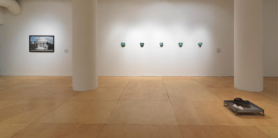 An installation photograph of a white wall in a gallery space. On the left side of the wall, a photograph of a federal building with white columns is mounted at eye-level. In the center of the wall, five green circular objects are situated in a horizontal line. Each object is nearly identical to the rest. There is about a foot of space between each green object.