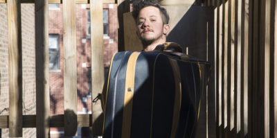 A photograph of a middle-aged person sitting inside a dark blue luggage bag. Their body is placed inside the bag, with their head uncovered. A dark, diagonal shadow crosses the photograph from the top right corner towards the bottom left corner. Behind the person inside the luggage is a wooden fence.