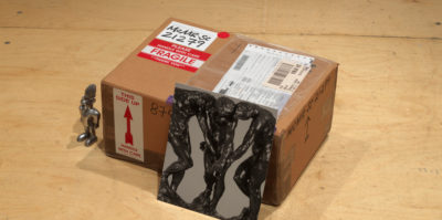 A photograph of a small cardboard shipping box. The box is wrapped with a shipping label.Placed against the box is an image of three sculptures of male figures. Sitting next to the box is a small, metal figurine.