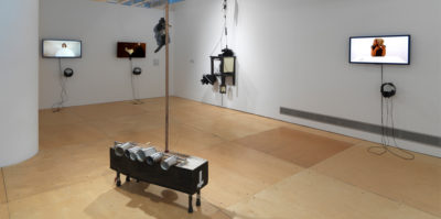 An installation photograph of a room in the gallery space. On the left wall, two television monitors are mounted on the wall. The monitor on the right is an image of a person with a white circular object, covering their body. The monitor on the right is of a person's foot in a dark room. Mounted on the right wall is an additional monitor and a square, three dimensional sculpture with four pegs. The monitor is paused on a film still of a middle-aged person sitting at a white table. In the center of the space, there is a rectangular, three dimensional object with six aluminum cans attached to the side.