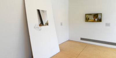 An installation photograph of two walls meeting in the corner of the gallery. On the left wall, a large, thick, white sheet leans against the wall. Centered in the middle of the sheet is a printed photograph of the same sheet. On the right wall, there is a framed, landscape style photograph. In the photograph there is a group of five people standing inside a room.