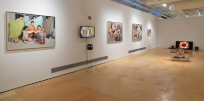 An installation photograph of the gallery wall. Starting from the farthest left, approximately a three by two foot photograph is mounted on the wall. The photograph is of four people sitting in a circle with their hands together. Three people in the background are standing, each holding a sheet. The second image is of a film still from a video on a television monitor, mounted on the wall. The next two photographs are also about three by two feet large. Both photographs are of a group of people sitting together. On the far end, a television monitor is placed on the ground with a star-like orange image paused on a film still.