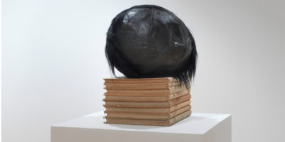 A photograph of a sculpture on a white podium. Eight, medium sized, light brown books are stacked vertically. Sitting on the stack of books is a black, circular object about the size of basketball.The circular object has black hair attached to its back side.