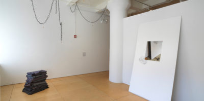 An installation photograph of the corner of a gallery space. On the right wall, a large, thick, white sheet leans against the wall. Centered in the middle of the sheet is a printed photograph of the sheet. In the left side of the space, on the floor, a vertical stack of black textiles are folded in squares and placed on top of one another. The size of the folded textiles from the floor is about two feet high.