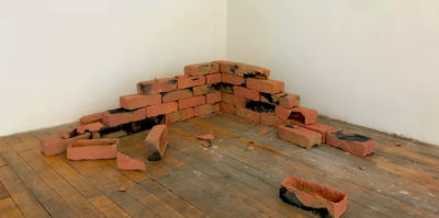 A photograph of a brick sculpture in the corner of the gallery. The brick layers form together to look like it was pulled from a structure’s corner, as the shape extends in opposite directions, fewer bricks are stacked. The bricks are orange in color, with stains of black and grey.