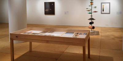 A installation photograph of a wooden and glass vitrine with documents and photographs inside. Behind the virtine, is a tall standing sculpture in the middle of the room consisting of a steel rod protruding from a large limestone base. Thread through the bottom of the rod directly above the limestone is a fragment of drainage tile. Above this, an assortment of green metal plates extends upwards. Against the wall that is behind a sculpture, are two different two-dimensional framed prints, about six feet apart from one another.