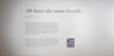 An installation photograph of the introductory paragraph text for the exhibition printed on the wall. The entire text is about 3 feet long, printed in a light grey font. The title is larger than the rest of the text which reads, “All have the same breath”.