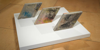 Three rhombus shaped concrete slabs parallel to each other resting on a white square platform. Upon the surface of the slabs there are oil pastel rubbings in various colors, the texture resembling crayons. (sky blue, hot pink, orange, and avocado green)