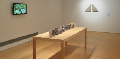 An installation photograph of three objects in the gallery. Displayed along a table is an accordion book with one whole side being a panorama of a garden, while the opposing side an outline of the geographic lands of planet Earth..On the wall adjacent to the accordion, is a video that is a documentation of moving plants accompanied by the sounds of wildlife. On the back centered wall there are two back to back, white, right angle triangle sculptures are positioned on the wall.