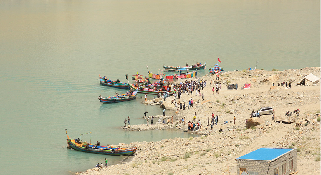 A photograph taken from above of people gathering on a beach. Six smaller boats, painted with bright colors, are lined up on the shore line.