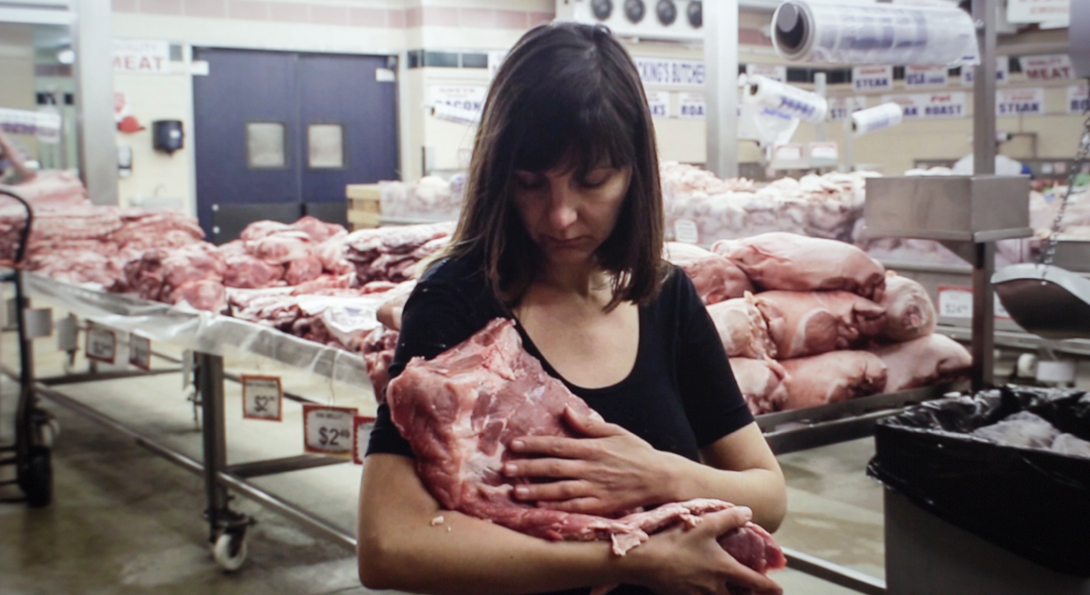 A photograph of a middle aged person holding a large, square piece of raw meat. They are holding it in their left arm, and their right arm is placed on the front side. The person is standing inside a meat producing facility. Behind them is a large table of stacked, multiple square pieces of raw meat. Another person, in a white sanitary suit is working on the table behind them.