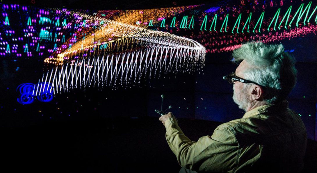 A photograph of a person with short gray hair. They are pointing a device to a screen that is wrapped around an entire wall, in a dark room. Multiple colors of shapes, lines and zig zags are pictured on the screen.