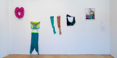 An installation photograph of five different materials mounted to the white gallery wall. The first material on the far left is of a bright pink cloth, weaved together in a circle. The second material to the right is a two-piece outfit with a blue mermaid tail. The top half of the two piece is a green and black and white checkered material. The third material are two full-sleeve gloves. One glove is teal and the other glove is a burnt, sparkling orange. The fourth material is in the shape of a blue purse, connected by two silver rings. The fifth and final piece to the right is a framed photograph of a person wearing the following materials that are mounted on the wall.