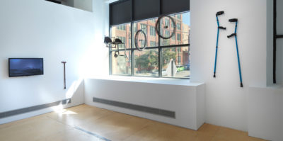 An installation photograph of two gallery walls. The gallery wall on the right has three windows. Hanging by an invisible wire in front of the window are two wheels and a chair piece. Next to the window is a mounted pair of blue crutches. On the wall left to the window is a mounted television monitor of a dark blue image. To the right is a mounted walking cane.