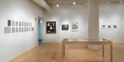 An installation photograph of a large coroner of the gallery space. On the right wall, six images are mounted on the wall. A large circular sits in the center, with three of the images on its left, and three on its right. On the other left wall, a series of unframed papers attached to the wall in two rows. On the floor in front of the two walls, a large, six foot long wooden vitrine is placed, showing multiple documents.