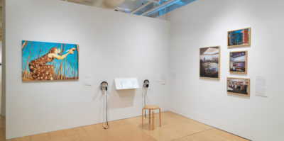 An installation photograph of the corner gallery space. The corner meets in the center of the photograph. The wall to the left has a large painting of a person painted in orange tones, with a blue background. Two pairs of headphones and a podium with documents is situated to the right side of the painting. On the right wall, there are four, wooden framed industrial photographs, mounted in a collage design.