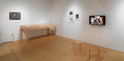 An installation photograph of the gallery. On the left wall, a small 12x16 image is framed and mounted. Against the left wall is a six foot long wooden vitine showcasing multiple documents. On the right wall, a tryptic of three 10x12 images with white frames are mounted. To the right of the tryptic is a television screen. The video still has three middle-aged people in conversation.