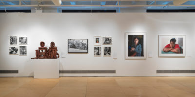 An installation photograph of seven artworks on a gallery wall. Starting from the far left side object, there are four small, black and white photographs in a white frame. The four photographs are aligned in a square formation. The second object is a brown clay sculpture of a person sitting down. The third object is an 11x17 black and white photograph of multiple people in a field. The fourth object is a series of four photographs aligned in a square formation. The top left photograph is in color, and the three additional photographs are in black and white. The fifth object is a large, framed, digital, portrait photograph of a younger-aged person. The sixth object is an additional large, framed, digital, portrait photograph of a younger-aged person by the same artist.