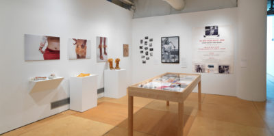 An installation photograph of the gallery space. On the left wall, three digital photographs of clothing are pinned to the wall. Below each photograph is a pedestal where the particular clothing piece is placed. On the right wall, a dozen small black and white photographs are placed on the wall in a collage formation. Next to this formation is a large, 2x3 foot, black and white photograph of a middle-aged person speaking into a microphone, on the right of this photograph is an even larger print of a poster with black and red text. In the open space between the two gallery walls is a wooden and glass vitrine where multiple documents are showcased.