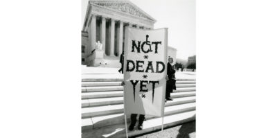 A black and white photograph of a person standing outside on the steps of a federal building. The person is holding a sign that is as large as their body. The sign writes, “NOT DEAD YET”. In the “O” letter of “NOT”, the “O” is designed to look like the accessible wheelchair symbol.