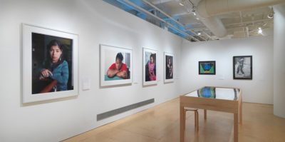 An installation photograph of the gallery space. In the center of the photograph, the wall on the left and right sides meet in the corner. On the left wall, four, large, white framed portraits are mounted. Each portrait is a portrait style photograph of a younger-aged person looking at the camera. On the right wall, two images are mounted on the wall in black frames. The image on the left has a blue shape in the middle with a black background and light green sides. The image on the right is a black and white drawing of a person standing. Centered on the gallery wooden floor is a large wooden and glass vitrine with documents inside.