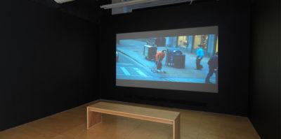 An installation photograph of the film room in the gallery space. The walls are black with a wooden bench in the center. Projected to the wall is a film still of a middle-aged person on a skateboard. They are skating on the sidewalk, wearing a red sweater, and two people are walking by them.