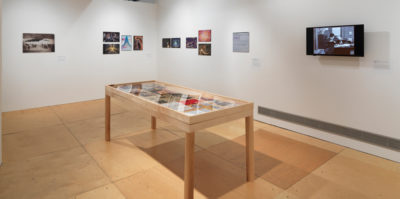 An installation photograph of the main gallery space. A large, seven foot long, wooden and glass vitrine is centered in the middle of the photograph. Inside the vitrine is a variation of photographs and documents. On the walls surrounding the vitrine are multiple, unframed images. On the wall to the right of the virtine is a modern-day television monitor with a black and white film still.