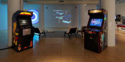 An installation photograph of the center of the gallery space. The left and right sides each have two ceiling length, white pillars, all together creating a square structure. Situated against the front two, left and right pillars there is each one video game console. Behind them is a grey rug with three white lounge chairs. On the back wall, a still from a projected video is displayed. The video still is of two middle aged people sitting next to eachother.