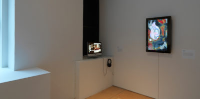 An installation photograph of the corner of the gallery space. On the left side of the corner, A small Mac monitor screen is playing a video. On the right wall of the corner, there is a television monitor positioned vertically on its side, with an abstracted blue, red, and white fluid design of different circular shapes.