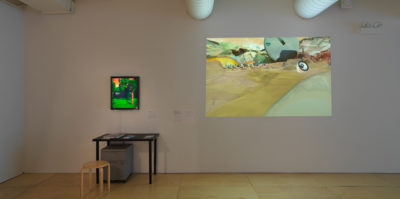 An installation photograph of the gallery wall. On the left side, a television monitor is mounted and is about the size of a 17x20 inch painting. The moniter has a saturated image of a tree in a park on its screen. Below the monitor, a metal black desk is situated against the wall with two books and a document. Underneath the desk is a two-door, metal filing cabinet. Parallel to this installation is a large, 4x6 foot projected image. The image is of a mountain landscape above a sand dessert with hills.