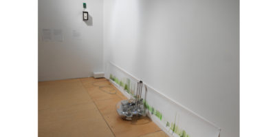 An installation photograph of two of the white gallery walls. On the bottom right wall, a long, two foot high print is displayed, touching the ground. In the center of the print, sitting against the wall, is a glass object with computer system objects inside. On the left wall, three white labels are placed in the center.