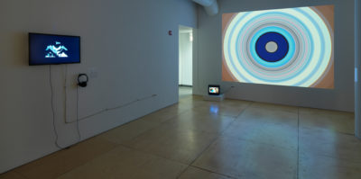 An installation photograph of the exhibition. The space lighting is darker. On the left wall, there is a flat-screen television with a film still of a black background and a blue design. A pair of headphones is attached to this screen for listening. On the wall in the center of the photograph, a large image of multiple circles is projected onto the white gallery wall. The projection size covers the entire gallery wall.