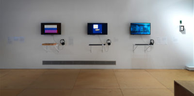 An installation photograph of the exhibition. This photograph is of one of the gallery walls. Mounted on the wall is three television monitors with headphones attached. Mounted on the wall below the televisions are individual shelves with square objects placed on top.