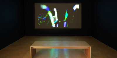 An installation photograph of the film screening room in the exhibition. The film is projected against the wall. The scene in the film still is of a middle-aged person that is pointing to an abstracted design. The film still is highly contrasted, with dark shadows. The abstracted light design is of three large, bright lines. To the left of the lines is a blue, green and red rainbow-like gradient.