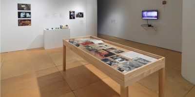 An installation photograph of the main gallery space. A large, seven foot long, wooden and glass vitrine is centered in the middle of the photograph. Inside the vitrine is a variation of photographs and documents. On the wall to the right of the virtine is a television monitor. On the monitor's screen is a blurred circular image in pink and blue tones. The wall on the left of the vitrine has five, unframed photographs positioned on the wall.