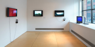 An installation photograph of the side gallery space. The right wall has a large window letting in natural sunlight. On the platform below the window, a small, 90’s style television is situated in the corner with a blue screen. The center wall has two, modern day television monitors of green and blue landscapes on the screens with headphones attached. The left wall also has a modern day television monitor mounted in the center. A pair of headphones is attached to the monitor. The screen has a red maroon color.