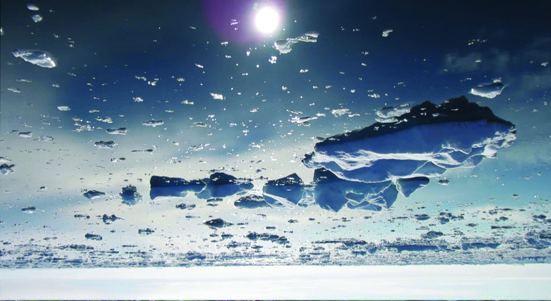 An upside-down photograph of melting glaciers in water. A white, circular glare from the sun is in the top center.