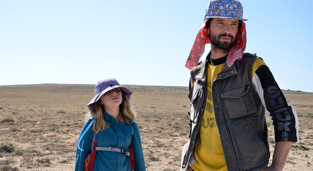 A photograph of two people standing in an empty, dry field. The person on the right is a middle-aged person wearing a yellow shirt. The person on the left is a younger-aged person in a blue jacket.