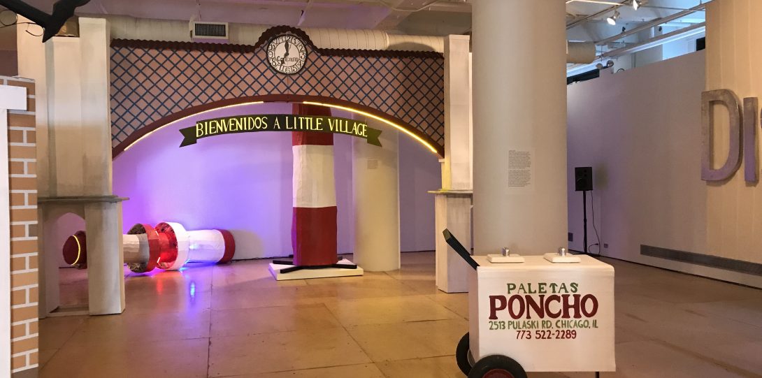 An installation photograph of The Last Judgment. This photograph is taken of the main gallery, in the center a large, maroon and brown arch sculpture that reaches from the floor to the ceiling. A white clock is centered in the top-middle point of the arch, below a ribbon design reads “Bienvenidos A Little Village”. In front of the arch, another sculpture, replicating a paletas cart sits on the right side of the photograph. The white paletas cart with red and green text and reads “Poncho, 2513 Pulaski RD, Chicago, IL”