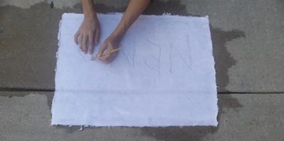 A digital photograph taken above of a white sheet of paper onto a sidewalk. The camera angle is pointing down, and the paper is in the center of the photograph. Wet marks from the paper sink into the cement, creating a darker, water stain. Two hands with a yellow pencil are extending out of the top center of the frame, writing on the white sheet of paper material.