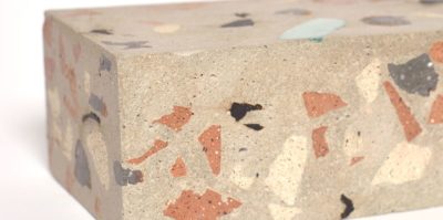 A digital photograph of a corner of a brick sculpture. This photo, taken very close to the sculpture, has a small amount of white background space in the top left corner. On the brick sculpture, marks in earth tones of brown, orange, beige, and black are painted in small, and are painted in various shapes and sizes.