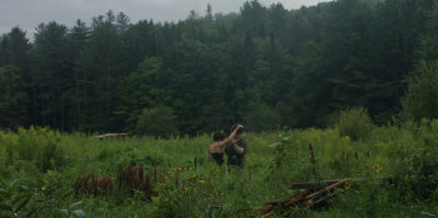 This film still is captured from the moving image piece. In the still, a moment between two middle aged people pauses while standing in a green, tall grass field. Behind them, a landscape of green, tall forest trees extend from the right to left center sides of the film still. One of the people is extending their right arm upward, and the second person, facing them, is holding that extended arm.