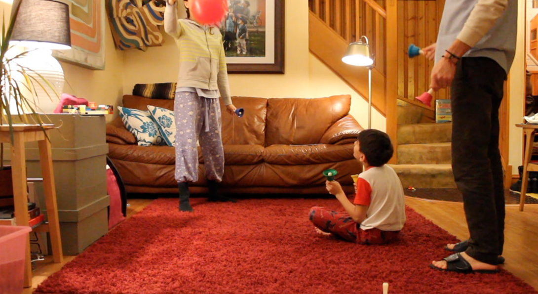 A photograph of a video still from Sensitive Equipment. The video still is in a living room setting. A young person is sitting on a red carpet on the floor. They are looking upwards at another young person, standing, holding a pink balloon. They are both wearing pajamas and engaging in front of a brown leather couch. Behind the couch, there are wooden stairs leading up to the top center of the photograph. On the right side of the photograph, the lower body of an older person is standing with toys in their hand.