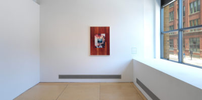 An installation photograph of a framed photograph centered on the gallery wall. The right wall has two windows, letting in natural light. The photograph is framed in a thin, white frame. The photograph begins with an image of dark wood as a border. Centered inside is another photograph of a framed image of the ocean. Behind the ocean image is a photograph of a burning bridge.