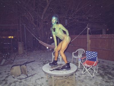 A photograph of a middle-aged person standing on a table in a backyard. The setting of the photograph is at night, while it is snowing. The middle-aged person is painted in full body paint. From their head to their feet, the body paint is in a gradient of blue, green, and orange. The person is wearing a gold necklace and black leather boots. Behind them, are plastic chairs, a fence, a hammock and a picnic chair. The picnic chair’s print material is the american flag symbol.